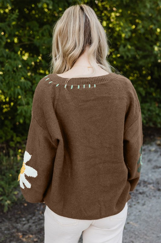 Person wearing a brown sweater with white accents, standing in front of green foliage.